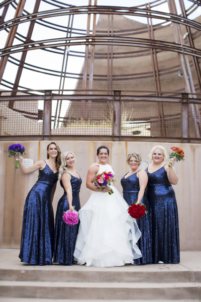 A bride in a white wedding dress poses with her bridesmaids. The bridesmaids are wearing sparkly blue dresses and each have a different colored floral bouquet. The bride has flowers from each of the other four bouquets in her bouquet. They are standing in front of a concrete and steel beam sculpture.