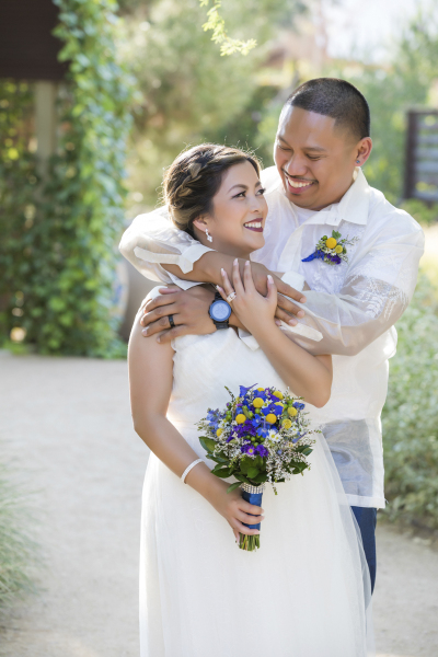 A bride and groom embrace as they pose for pictures on their wedding day at Springs Preserve in Las Vegas. The smiling latino couple are each wearing white. She holds a bouquet of yellow and blue flowers and he wears a matching boutonniere. 