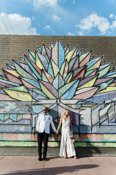 A bride and groom posing in front of a brick mural. They mural is painted with a colorful geometric crystal