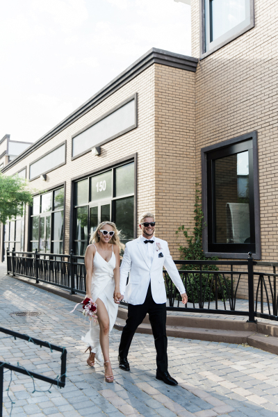 A bride and groom hold hands and walk through an outdoor brick-lined corridor. He wears a white tuxedo jacket and they both wear sunglasses.