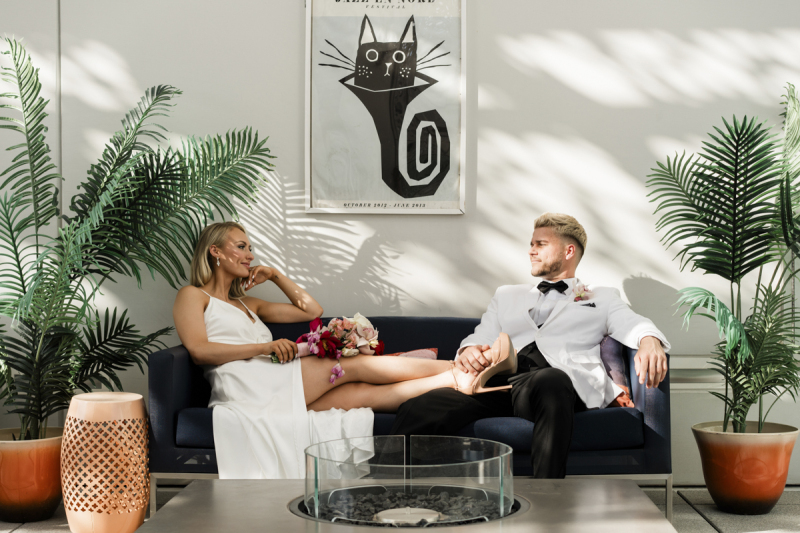A bride and groom sit on a blue couch in front of an unlit fire pit. She sits sideways with her legs up on his knee. He turns to smile at her. They are lit by dappled sunlight.