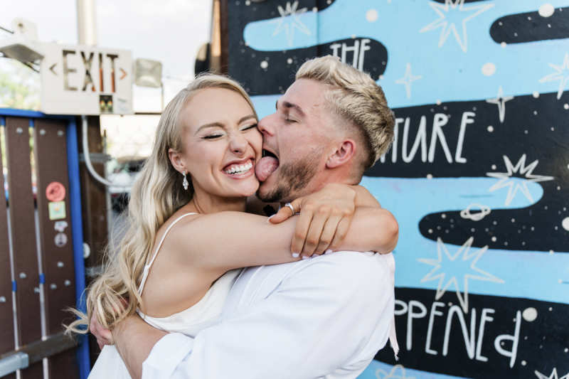 A bride and groom embrace. The bride smiles wide as the groom licks her face. They are being silly on their wedding day at the Beverly Theater in Las Vegas.