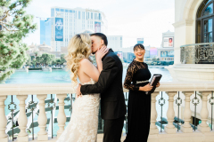 A bride and groom kiss on the balcony at the Bellagio as their officiant looks on and smiles.