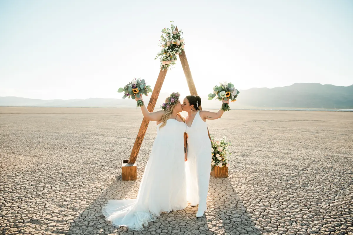 two brides stand in a dry lake bed and kiss as they hold their floral bouquets up.