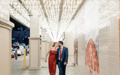 A couple in dressy clothes walks toward the camera leaning in for a kiss. They're outside in Las Vegas, walking under an awning covered in lights.