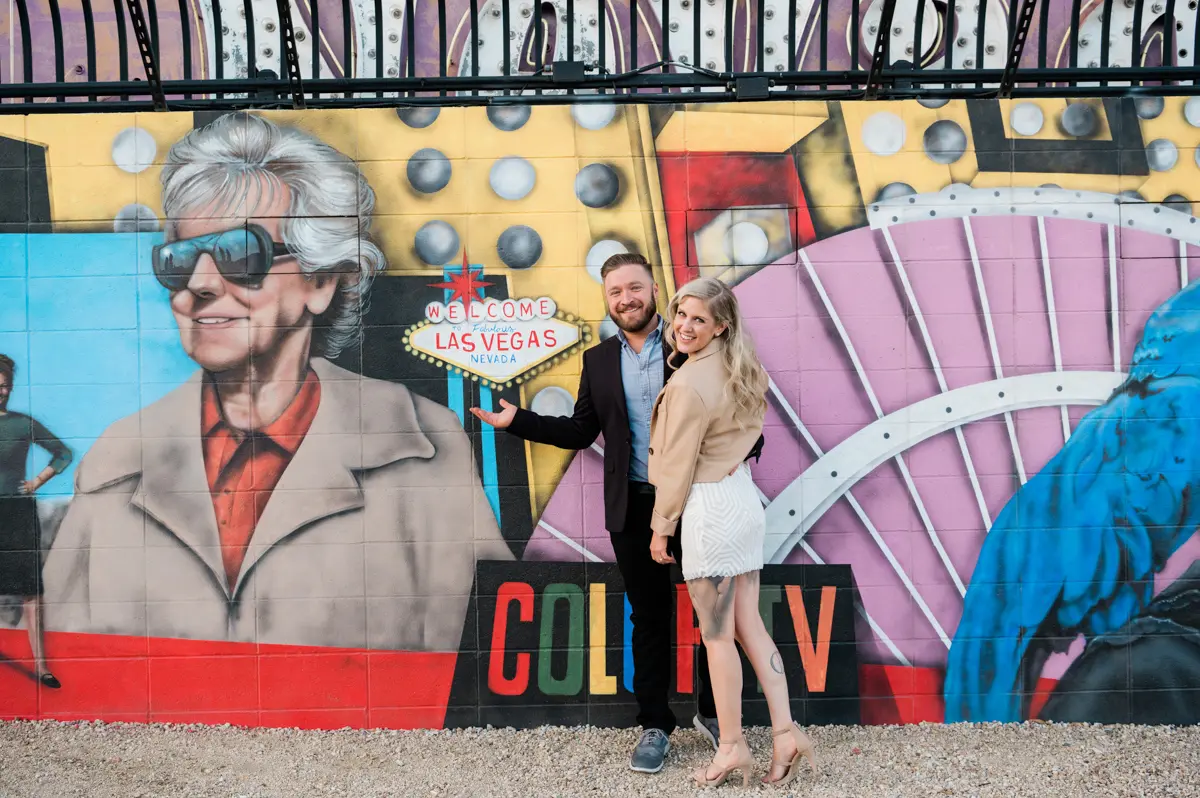 an engaged man and woman stand in front of a colorful mural with a las vegas theme, smiling at the camera.
