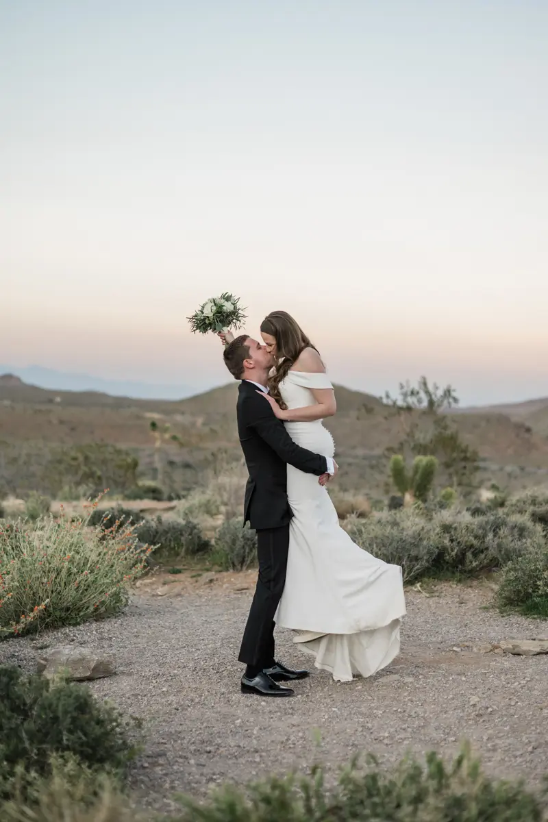 A bride and groom sharing a romantic kiss in a desert landscape during their las vegas wedding, with the groom lifting the bride into the air against a sunset sky.