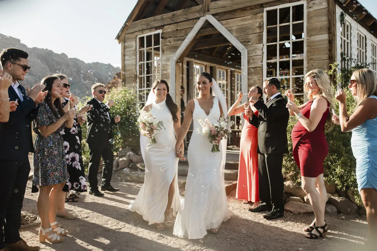 Two brides walking through a bubble exit outside a rustic wooden chapel for their Las Vegas wedding.
