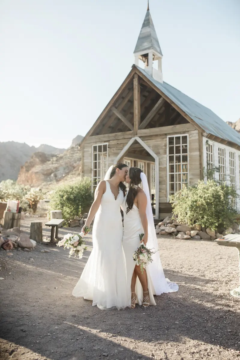Smiling bride and groom posing against desert red rock formations for their las vegas wedding. Two brides kissing in front of a rustic wooden chapel during their Las Vegas wedding.