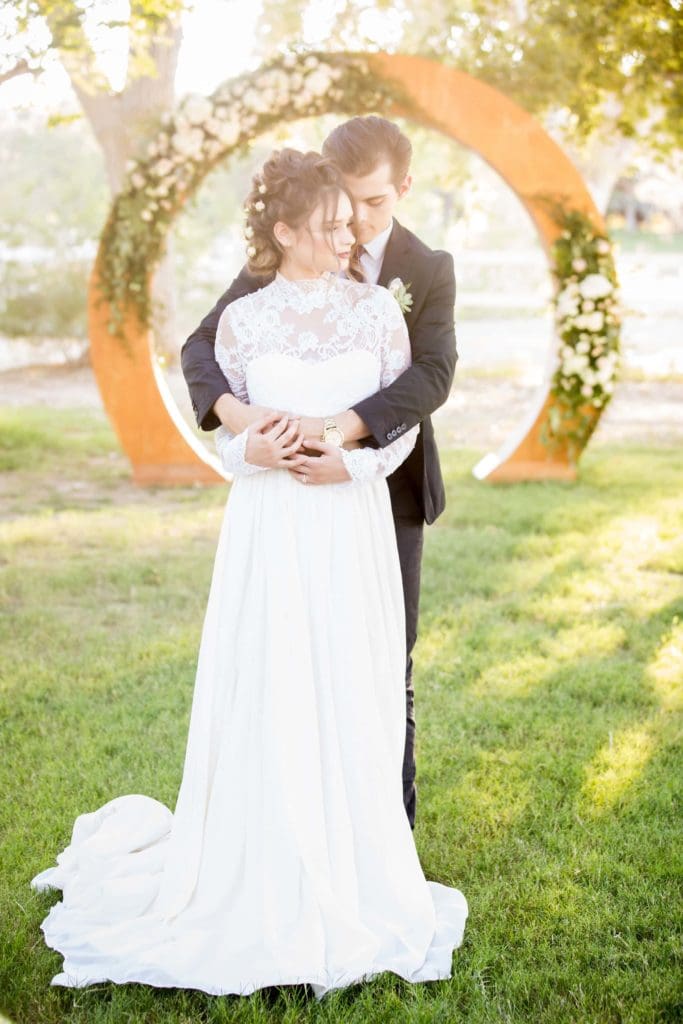 Groom with arms wrapped around bride in the park.