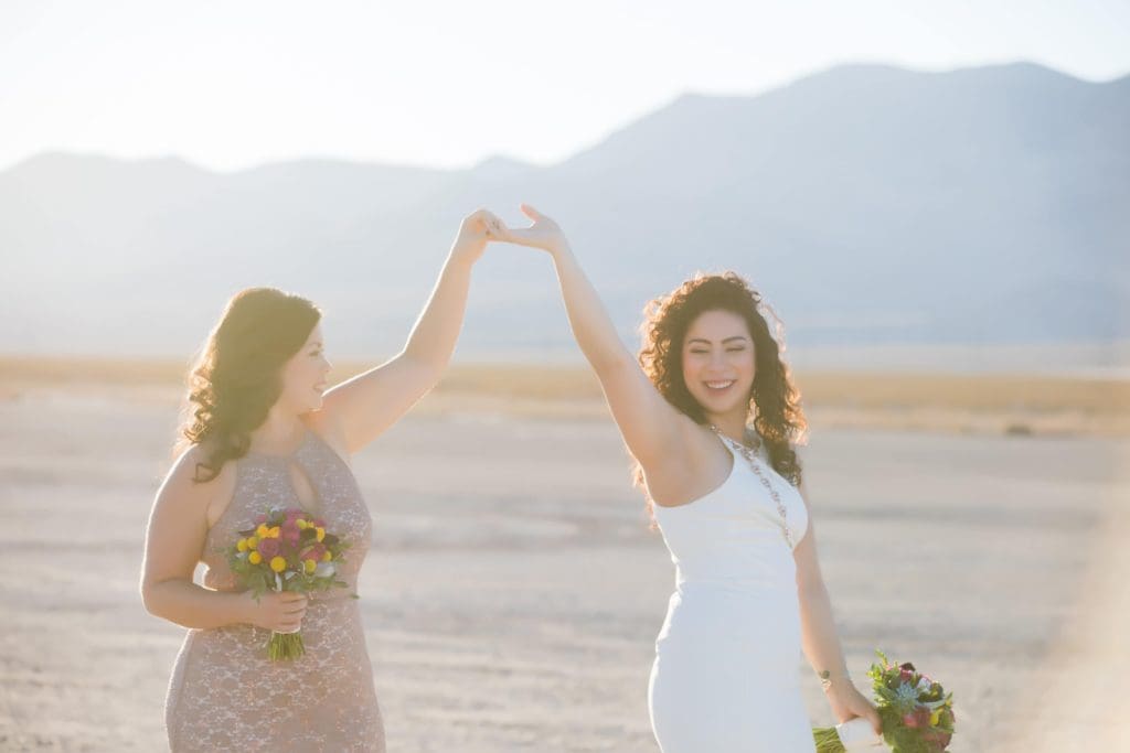 Two brides hold hands in the air in the Las Vegas desert.