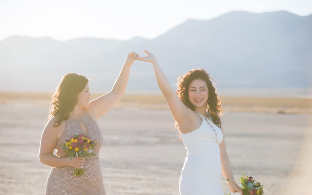 Two brides hold hands in the air in the Las Vegas desert.