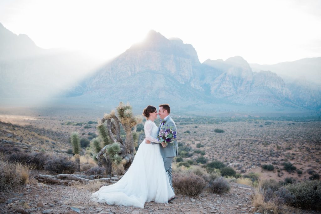 Newlywed couple standing face to face in front of a mountain with the sun shining from behind.
