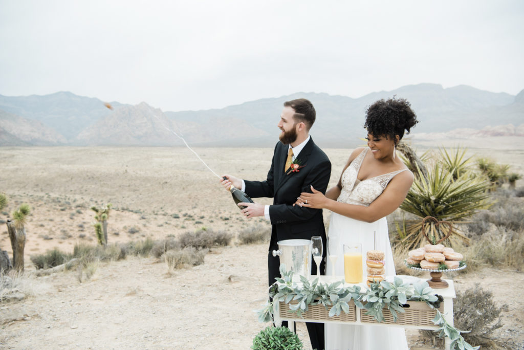 Smiling bride and groom, opening a bottle of champagne in the desert.