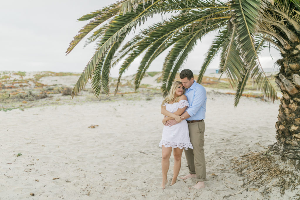 A bride and groom stand barefoot on the beach under a palm tree. He stands behind her with his arms around her wearing a blue button up and khakis, while she wears a short white dress with eyelet details.