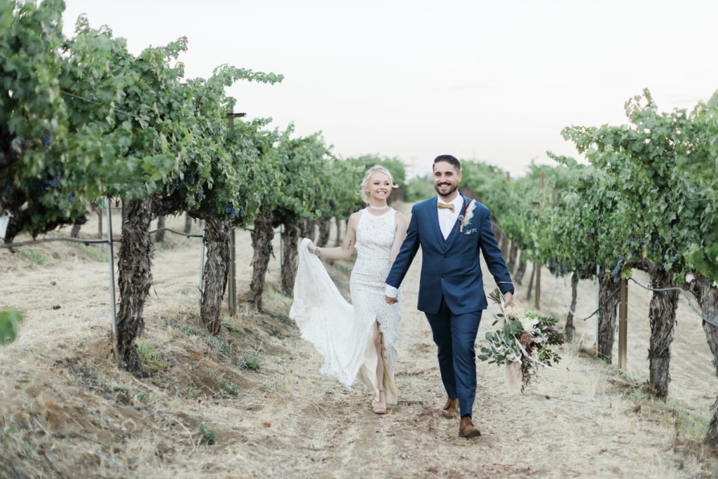A bride and groom smile and walk, holding hands, in the Wilson Creek Winery. She wears a high-necked sleeveless white dress with a front slit and he wears a blue suit with a bow tie.