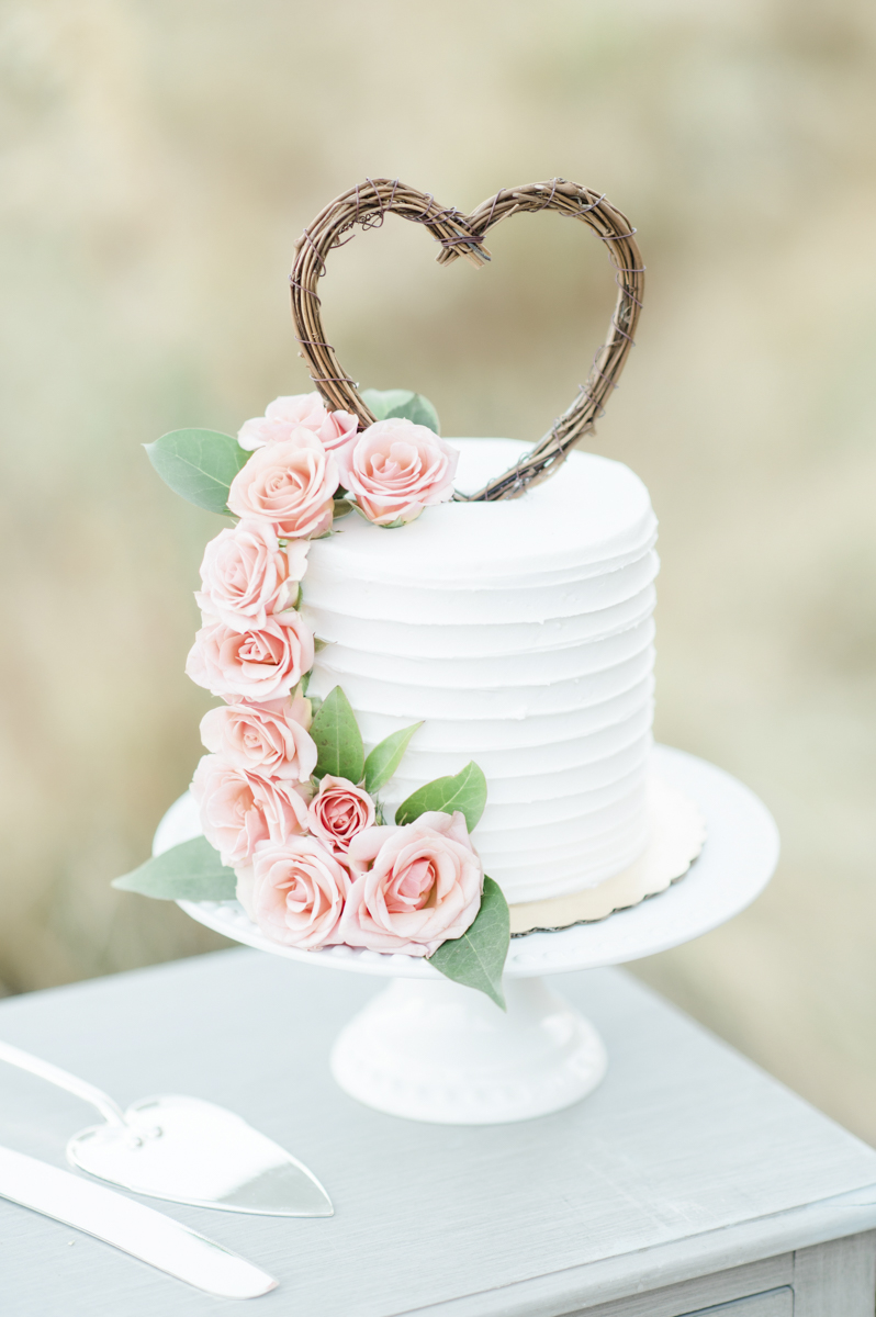 White wedding cake with heart topper and pink flowers along the side.