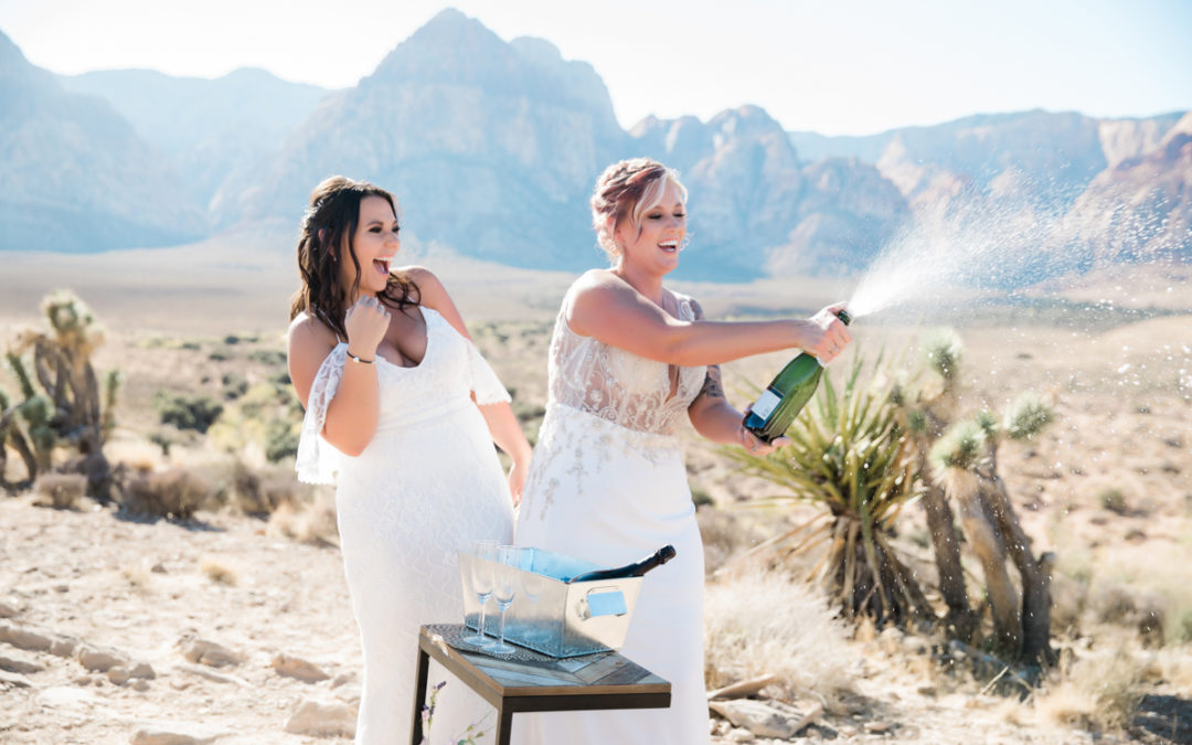 two happy brides in the desert with one popping a bottle of champagne