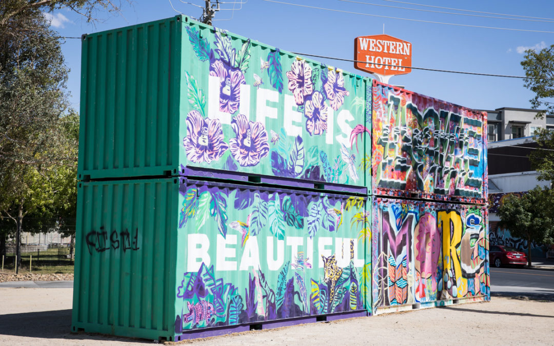 Two large cargo containers are stacked and tagged with grafitti. They read "Life Is Beautiful" in white letters on a mint green background and surrounded by purple flowers.