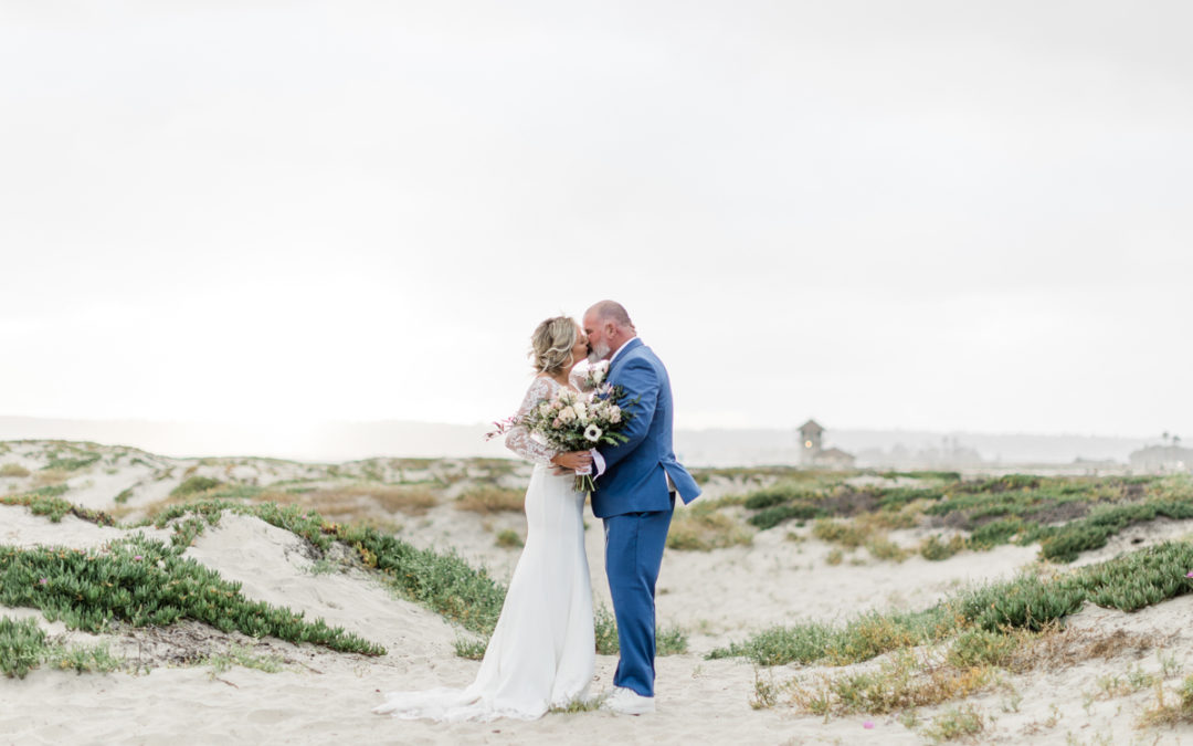A bride and groom kiss on Coronado Beach dunes. Bride is holding a big white and green bouquet.