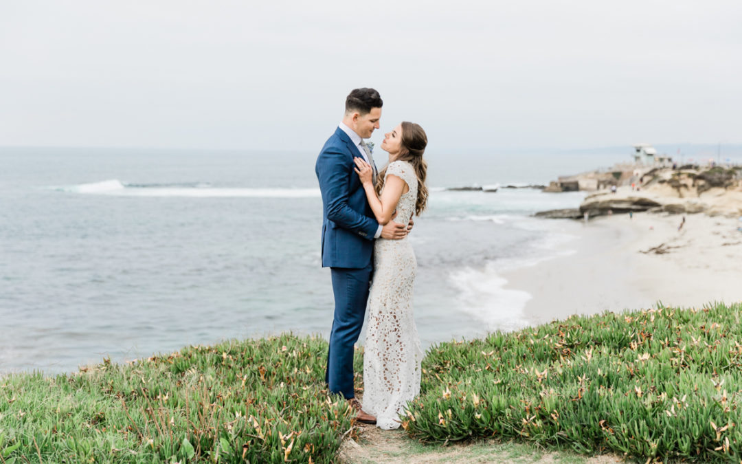 a groom and a bride embrace and look at each other as they stand on a grassy hill with the beach and ocean behind them.