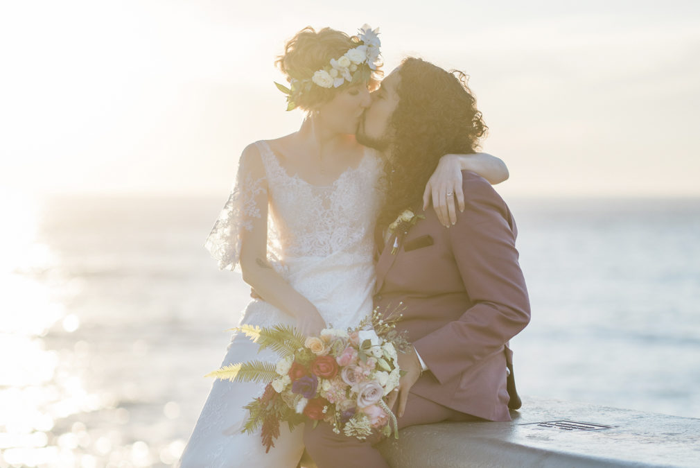 A bride wearing a floral crown sits on the lap of her groom with long hair as they kiss, with a large body of water behind them.