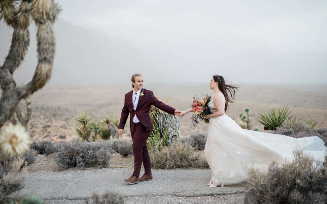 A groom in maroon walks to the left holding his bride's hand and looking back at her. They're in the desert and she's carrying a big bouquet and the wind is blowing the train of her white dress backward.