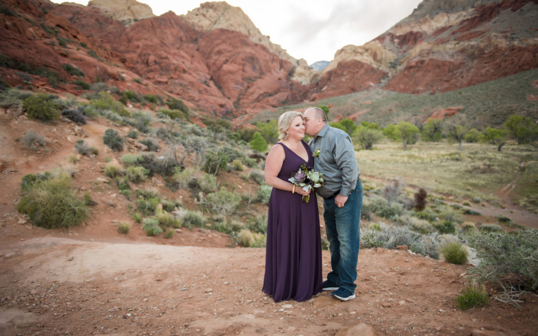 A husband kisses his wife on her cheek as they stand on a vista overlooking a valley and with mountains in the distance, after their vow renewal ceremony.
