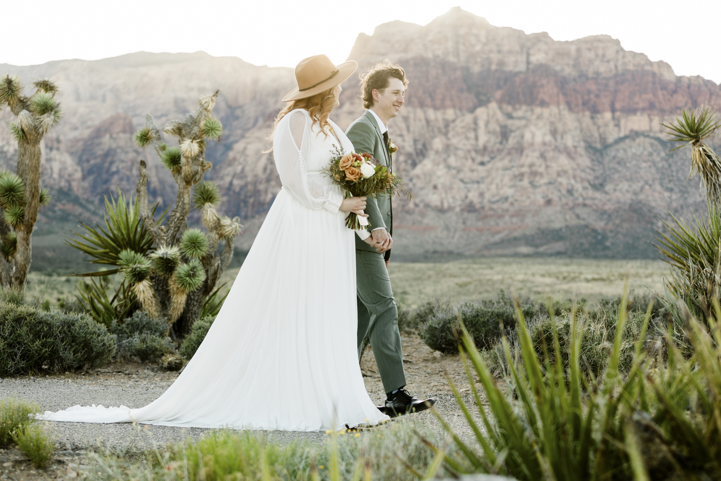 A couple walks hand-in-hand through a scenic desert landscape, framed by Joshua trees and rugged Red Rock Canyon mountains.