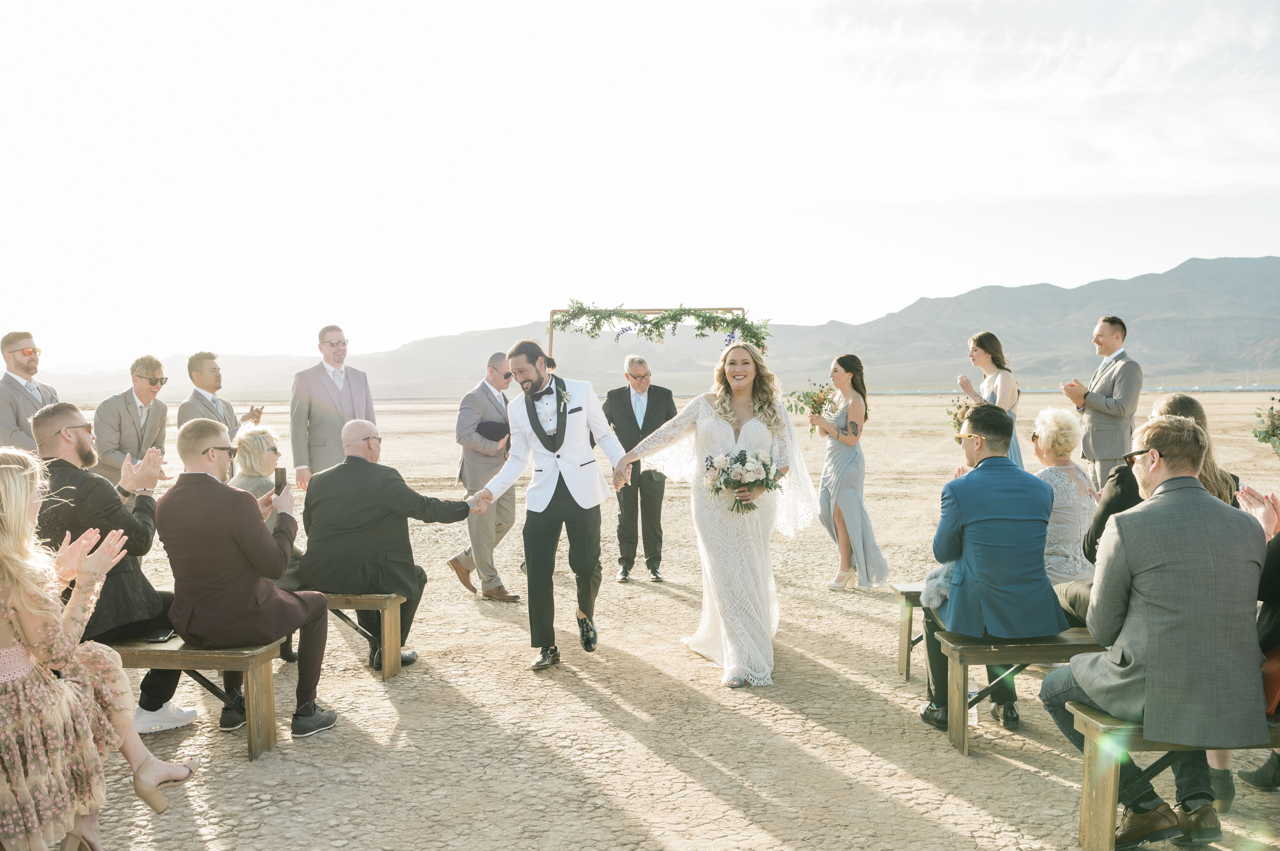 A joyful couple exits their intimate desert wedding ceremony while guests applaud enthusiastically.