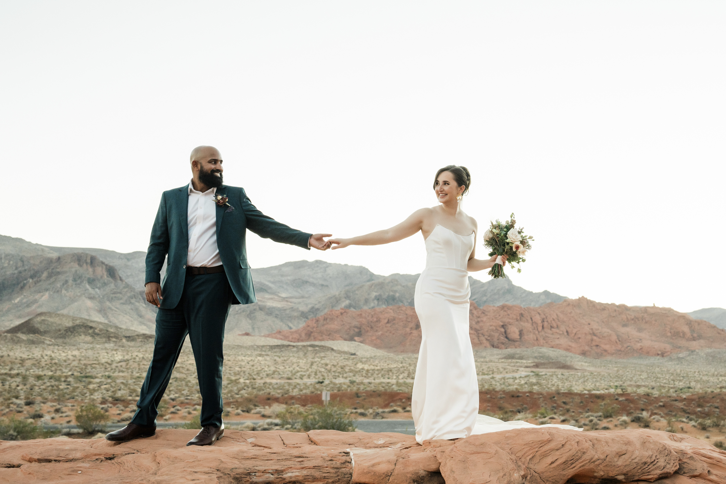 A newly engaged couple hold hands atop a red rock formation, framed by breathtaking desert and mountain views.